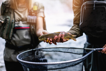 Young adult couple is fishing together on fast mountain river. They are holding a live trout before...