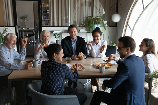 Smiling diverse employees sit in office have fun enjoy pizza on work lunch break together. Happy multiethnic colleagues coworkers laugh joke eating Italian fast food meal, drink coffee at workplace.