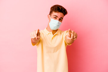 Young caucasian man wearing a protection for coronavirus isolated on pink background cheerful smiles pointing to front.