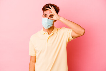 Young caucasian man wearing a protection for coronavirus isolated on pink background excited keeping ok gesture on eye.