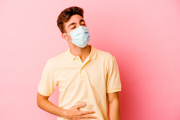 Young caucasian man wearing a protection for coronavirus isolated on pink background touches tummy, smiles gently, eating and satisfaction concept.