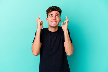 Young caucasian man isolated on blue background crossing fingers for having luck