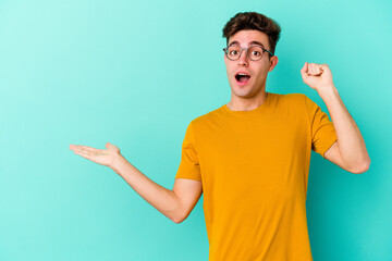 Fototapeta premium Young caucasian man isolated on blue background holds copy space on a palm, keep hand over cheek. Amazed and delighted.