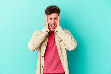 Young caucasian man isolated on blue background covering ears with hands trying not to hear too loud sound.