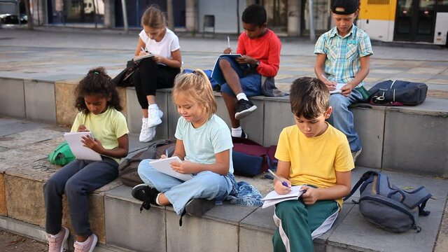 Multiethnic group of schoolchildren sitting with workbooks in schoolyard in warm autumn day. High quality FullHD footage