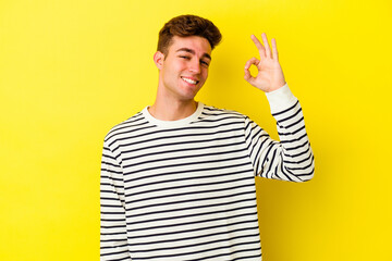 Young caucasian man isolated on yellow background winks an eye and holds an okay gesture with hand.