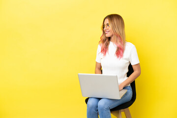 Young woman sitting on a chair with laptop over isolated yellow background looking side