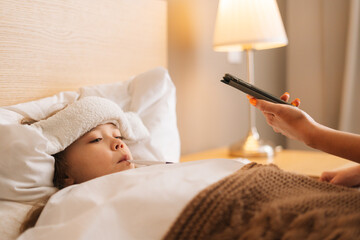 Close-up of worried mother sitting on bed beside sick daughter with high fever and calling on phone...