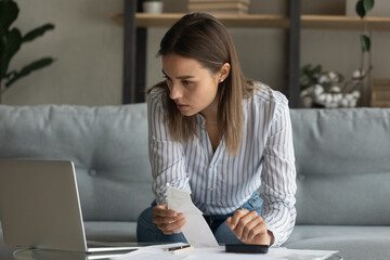 Close up serious woman calculating expenses, checking financial documents, domestic bills, sitting on couch at home, focused young female using laptop, looking at screen, managing planning budget