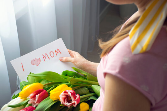 Mother holds in hands bouquet of colorful tulips and handwritten card with words I Love Mom drawn by child. Happy Mother`s Day or Happy Birthday greetings and congratulations to mommy.