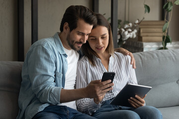 Close up young couple using gadgets together, sitting on couch, woman and man spending leisure time with smartphone and tablet, looking at phone screen, chatting or shopping online at home