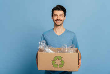 Smiling unshaved conscientious young man in blue t-shirt holds a cardboard box full of a plastic...