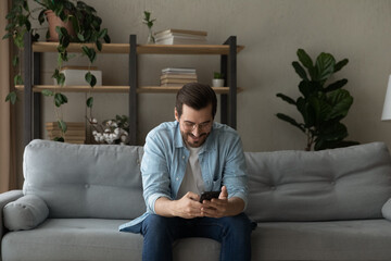 Obraz premium Smiling man wearing glasses having fun with phone at home, sitting on couch, looking at mobile device screen, chatting or shopping online, browsing apps, enjoying leisure time with smartphone