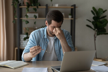 Tired businessman massaging nose bridge, feeling eyestrain, holding glasses, sitting at work desk with laptop, exhausted young man suffering from dry eye syndrome, feeling fatigue and dizziness