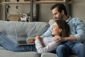 Side view close up smiling woman using laptop, lying on boyfriend laps, relaxing on couch at home, happy young couple looking at computer screen, shopping or chatting online, enjoying leisure time