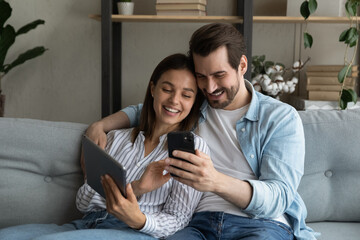 Close up happy young couple having fun with mobile devices together, using smartphone and tablet, smiling woman and man hugging, looking at phone screen, chatting or shopping online at home