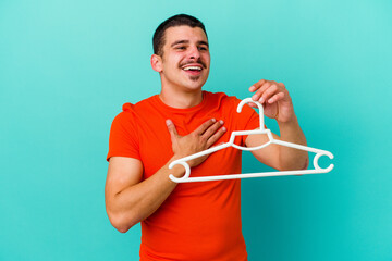Young caucasian man holding a hanger isolated on blue background laughs out loudly keeping hand on chest.