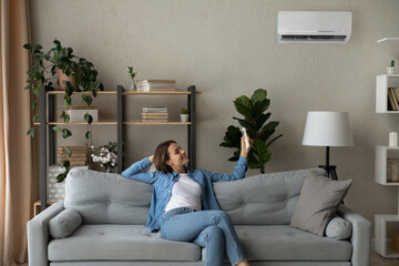 Smiling woman using air conditioner remote climate controller, switching setting comfortable temperature in modern living room, happy young female relaxing on cozy couch at home, enjoying fresh air