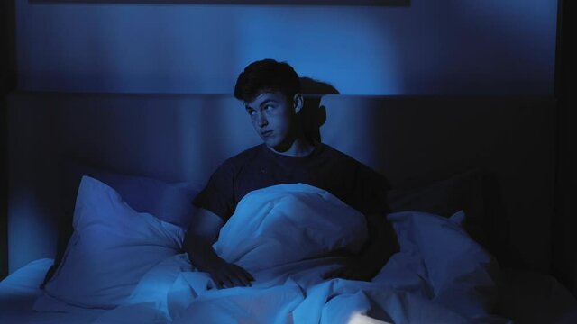 Night insomnia. Sleep disorder. Depression anxiety. Tired bored pensive guy cant rest awake late yawning sitting alone in bed at home in dark blue light.