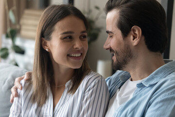 Close up happy wife and husband talking, enjoying tender moment, sitting on couch, hugging, smiling young couple chatting, expressing love and care, spending free time together, romantic date