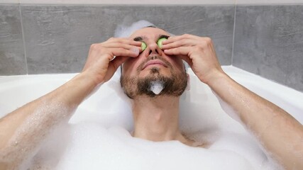 Handsome bearded guy relaxing inside bath with foam, applying cucumber eye mask. Spa treatment at home bathroom. Man wellness, body care and self love concept. - Powered by Adobe