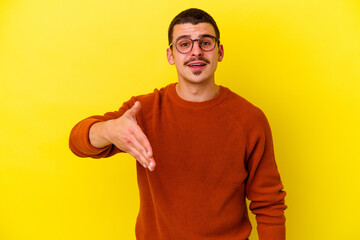 Naklejka premium Young caucasian cool man isolated on yellow background stretching hand at camera in greeting gesture.