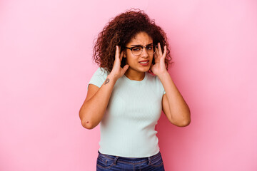 Young african american woman isolated on pink background covering ears with fingers, stressed and desperate by a loudly ambient.