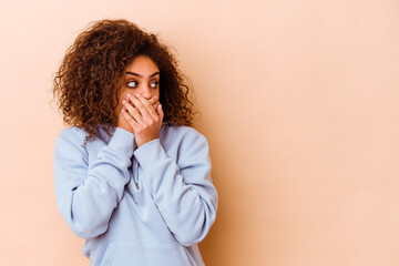 Young african american woman isolated on beige background thoughtful looking to a copy space covering mouth with hand.