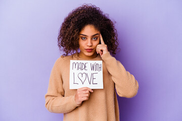 Young African American woman holding a Made with love placard isolated on purple background pointing temple with finger, thinking, focused on a task.