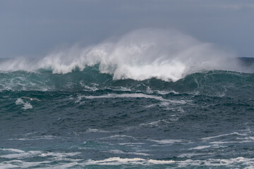 A long wave with a white spray coming over the top of the rip curl. It's a grey gloomy day with clouds in the sky. The water is vibrant teal green in colour. The light is going through the curl.