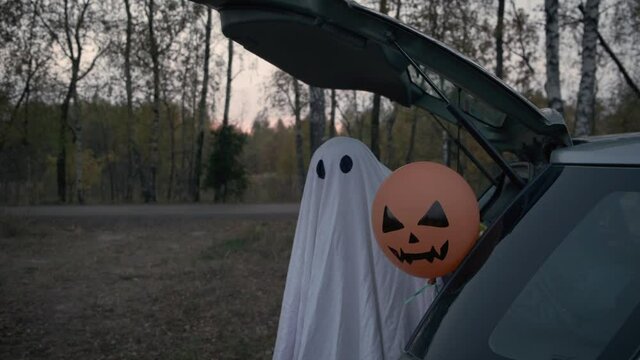Halloween concept. Cute little child in ghost costume holding scary face orange air balloon while sitting inside car trunk near twilight autumn forest road.