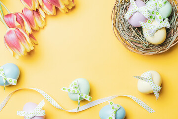 Easter basket bunny. Colourful egg with tape ribbon, spring tulips, feathers on pastel yellow background in Happy Easter decoration. Festive design in flat lay.