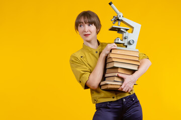 Woman with a book and a microscope in her hands. Microscope as a symbol of research. Portrait of a female explorer on a yellow background. Student wants to do research. She prepares for lab work