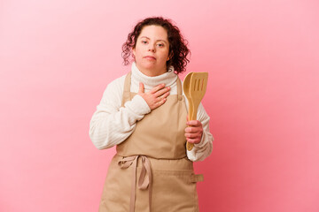 Woman with Down syndrome cooking at home isolated on pink background laughs out loudly keeping hand on chest.