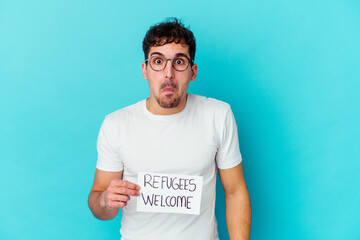 Young caucasian man holding a Refugees welcome placard isolated shrugs shoulders and open eyes confused.