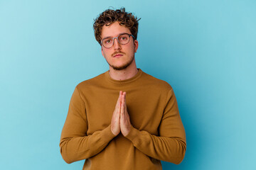 Young caucasian man wearing eyeglasses isolated on blue background praying, showing devotion, religious person looking for divine inspiration.