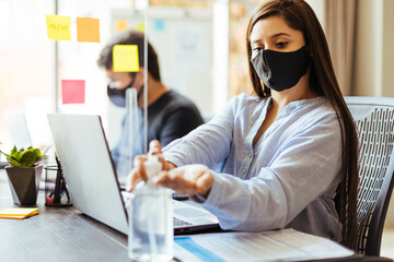 Business team wearing protective masks sitting at their desks separated by plexiglass dividers....