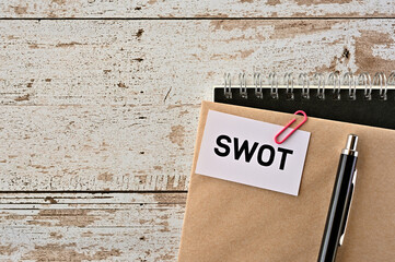 There is a piece of paper with "SWOT" written on it. It was an abbreviation for Strength, Weakness, Opportunity, Threat. It was on top of white damaged wood table with a envelope and a notebook.