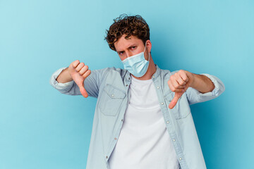 Young caucasian man wearing an antiviral mask isolated on blue background showing thumb down and expressing dislike.