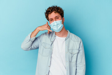 Young caucasian man wearing an antiviral mask isolated on blue background showing a mobile phone call gesture with fingers.