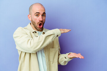 Young caucasian bald man isolated on purple background shocked and amazed holding a copy space between hands.