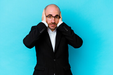 Young business caucasian bald man isolated on blue background covering ears with hands trying not to hear too loud sound.