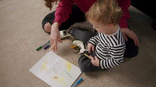 Happy young mother and little son are sitting on the floor, painting with colourful markers. Mom is teaching her child how to draw. Lots of colored felt pens and papers around. 