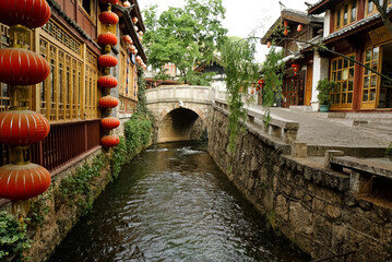 Fototapeta na wymiar Tile-roofed shops, restaurants, and guest houses front a stone-walled canal with arched stone bridge in the ancient town of Lijiang (Dayan), Yunnan Province, China.