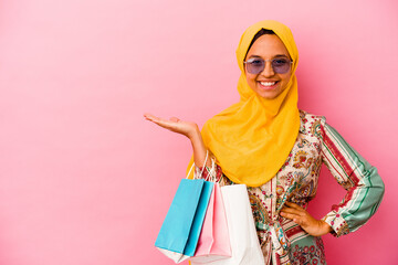 Young muslim woman shopping some clothes isolated on pink background showing a copy space on a palm and holding another hand on waist.