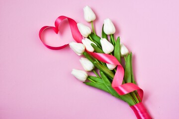 White tulips with pink ribbon in the form of a heart on a pink background.  Flat lay, copy space. Valentines Day and Womens Day, mother's day concept. 