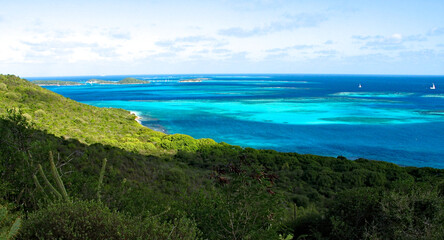 View from Mayreau towards Tobago Cays: exquisite blue lagoon, lush green vegetation, coral reef and charter boats sailing away from Tobago Cays islands.
