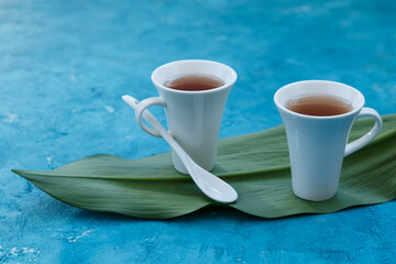 Two white cups with ceramic teaspoon with tea standing on a green leaf with blue background