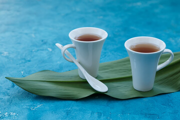Two white cups with ceramic teaspoon with tea standing on a green leaf with blue background