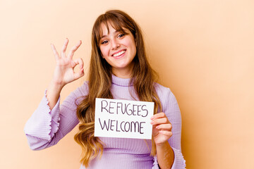 Young caucasian woman holding a Refugees welcome placard isolated cheerful and confident showing ok...